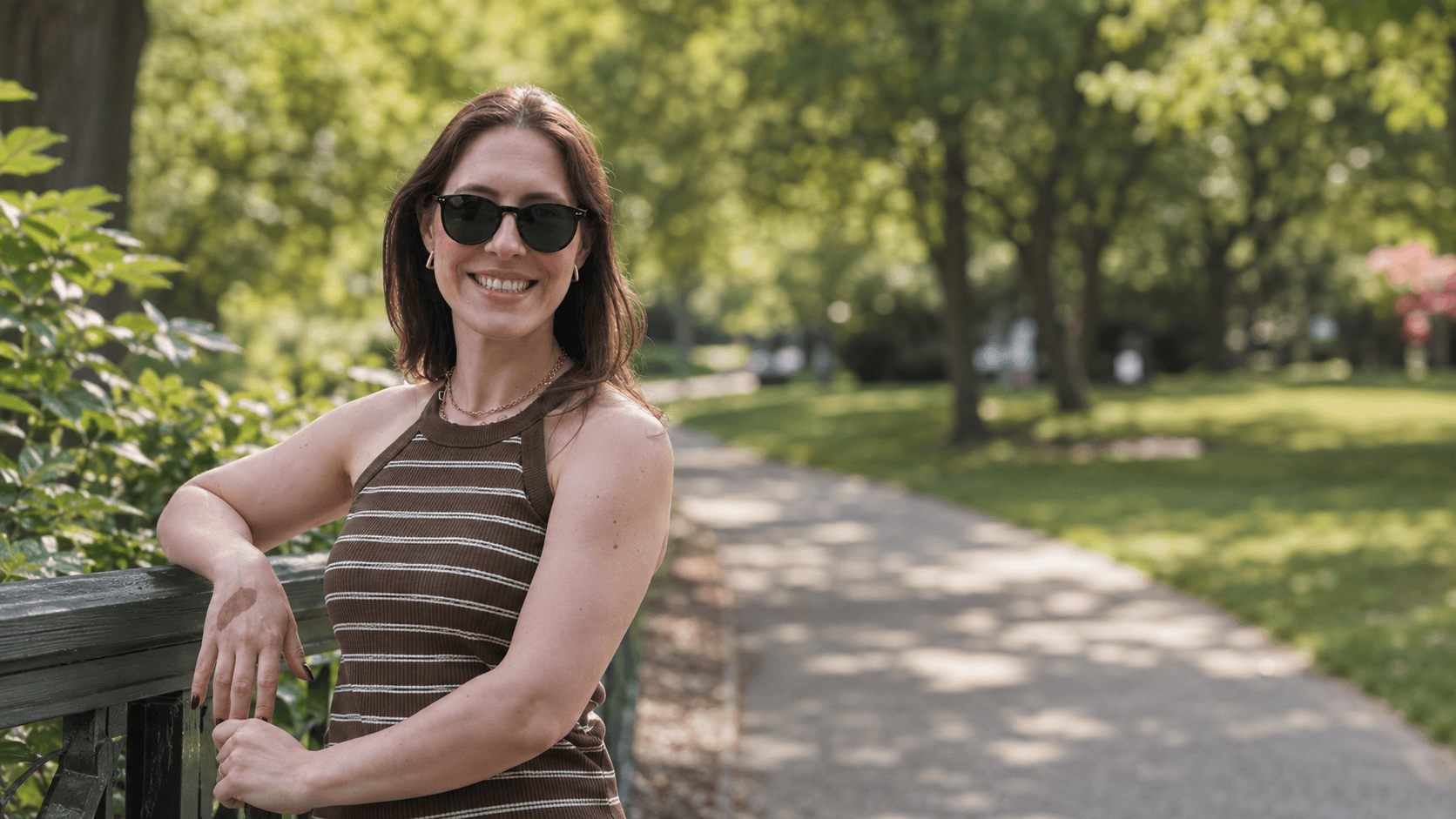 Mulher sorrindo, apoiada em um parapeito, durante uma caminhada em um parque